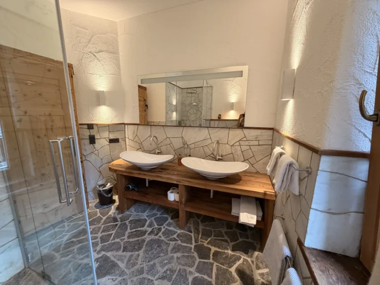 Badezimmer des Studios Wasserfall mit Glasdusche, Naturstein und zwei Waschbecken auf einer Holzablage